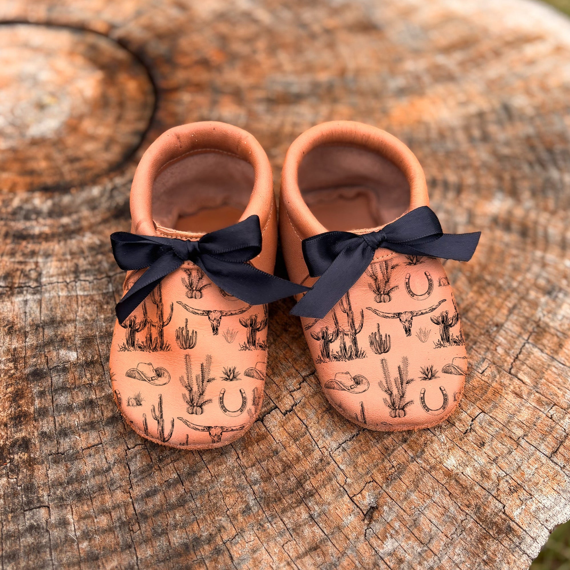 Pair of baby shoes with cactus pattern and black bows on a wooden surface.